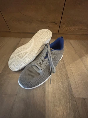 Grå sneakers från Kappa - Snygga grå sneakers från Kappa med vit sula och blå insida. Ovandelen är i meshmaterial för bra ventilation och skorna har klassisk snörning. Perfekta för dig som vill ha en stilren och sportig look.