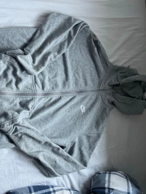 Grå zip hoodie från Nike - Säljer en grå zip hoodie från Nike med vit logga på bröstet. Hoodien har dragkedja hela vägen, två fickor fram och en klassisk huva med dragsko. Tillverkad i mjukt bomullsmaterial som är skönt att ha på sig. Perfekt för chill dagar eller till träningen.