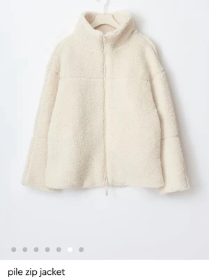 Krämvit teddyjacka med dragkedja - Mysig teddyjacka i krämvit färg med hög krage och hel dragkedja framtill. Jackan har en avslappnad passform och är tillverkad i fluffigt pile-material som ger en trendig och varm look. Perfekt för lager-på-lager-stil under kyliga dagar.