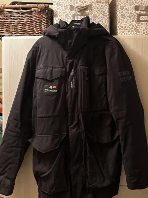 Sail racing jacka  - Snygg svart parkasjacka från sail racing  med huva, flera stora fickor och coola patchdetaljer på bröstet och ärmen. Jackan har dragkedja och vindslå framtill, perfekt för kalla dagar. Klassisk och stilren design med sportig känsla.