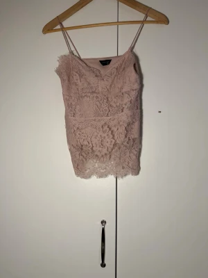 Rosa spetslinne från Vero Moda - Säljer ett ljusrosa linnetopp från Vero Moda med tunna axelband och vacker spetsdetalj över hela toppen. Perfekt för att lyfta vilken outfit som helst med sin romantiska känsla och feminina look.