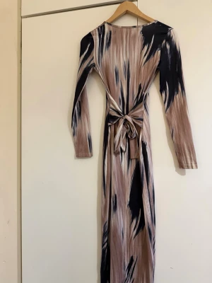 Mönstrad långklänning med knytband - Långklänning med beige, svart och vit abstrakt mönster. Klänningen har långa ärmar, v-ringning och ett brett knytband i midjan som markerar siluetten. Tillverkad i mjukt, stretchigt material som ger en bekväm passform. Går att knytas fram eller bak.