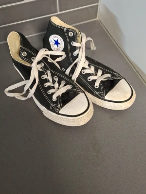 Svarta Converse Chuck Taylor High - Klassiska svarta Converse Chuck Taylor All Star high-tops med vit tåhätta, vita snören och kontrastsömmar. Skorna har den ikoniska loggan på sidan och är tillverkade i canvas med gummisula. Perfekta för en avslappnad och tidlös streetstil.