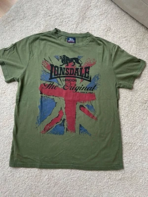 Grön Lonsdale t-shirt - Snygg grön t-shirt från Lonsdale med stort tryck av Union Jack och logga på framsidan. Klassisk passform och korta ärmar. Materialet är 100% bomull. Skriv vid minsta fundering, kan även ta fler bilder osv. Hör av dig! Skulle även vela säga att den är i nytt skick det finns inget tecken på användning. 