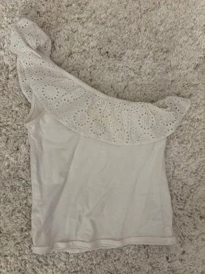 Vit oneshoulder topp med volang - Snygg vit oneshoulder topp med bred volang i broderad spets längs axeln. Toppen är ärmlös och har en feminin, somrig look. Perfekt att styla med jeans eller kjol för en trendig outfit. Toppen har en liten fläck på framsidan men inget som märks till tycker jag