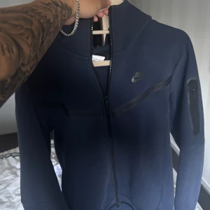 Nike Tech Fleece - Säljer en mörkblå hoodie från Nike med hel dragkedja och huva. Tröjan har svarta detaljer, en ficka med dragkedja på ärmen och Nike-logga på bröstet. Perfekt för chill eller träning, riktigt snygg och skön passform.