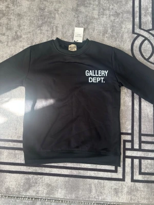 Svart sweatshirt från Gallery Dept. - Snygg svart sweatshirt från Gallery Dept. med vit logga tryckt på bröstet. Tröjan har rund halsringning och långa ärmar. Tillverkad i mjukt material som känns skönt mot huden. Perfekt för en clean och stilren look.