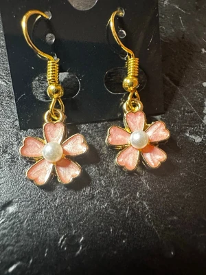 Blommiga örhängen med pärldetalj - Säljer ett par söta örhängen i form av blommor med rosa blad och guldfärgade kanter. I mitten sitter en liten vit pärla som ger extra söt touch. Krokarna är också guldfärgade och örhängena har en lekfull och trendig look.