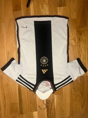Tysklands fotbollströja Adidas - Säljer en vit och svart Tysklands landslagströja från Adidas med guldiga detaljer och broderad logga på bröstet. Tröjan har klassiska Adidas-ränder på ärmarna och en snygg krage med röd och gul kant. Perfekt för fotbollsfans!