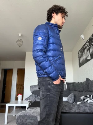 Moncler Agay Blå dunjacka (M) - Snygg blå dunjacka från Moncler med hög krage och två dragkedjeförsedda fickor framtill. Jackan har quiltad design och Moncler-logga på ärmen. Perfekt för kalla vinterdagar och ger en cool look.