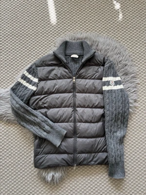 Grå cardiganjacka från Moncler (L) - Säljer en grå cardiganjacka från Moncler med quiltad front och stickade ärmar. Jackan har dragkedja, ribbad krage och två vita ränder på ärmarna samt Moncler-logga på vänster ärm. Perfekt för lager-på-lager och snygg till jeans.