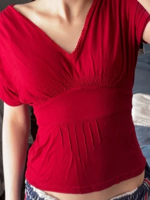 Röd v-ringad topp  - Snygg röd topp med v-ringning och smockdetaljer under bysten. Kortärmad och figurnära passform som framhäver midjan. Tunn och mjuk bomullskvalitet, perfekt för att matcha med jeans eller kjol. Diskret spetskant vid halsen ger en extra touch.