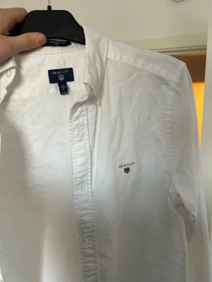 Vit skjorta från GANT med logga - Vit klassisk skjorta från GANT med broderad logga på bröstet. Skjortan har knappar hela vägen och en krage som ger en stilren look. Materialet är bomull och modellen är långärmad, perfekt för en clean och snygg outfit.