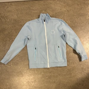 Ljusblå ziptröja från BOSS - Ljusblå ziptröja från BOSS med vit dragkedja och två fickor med dragkedja. .