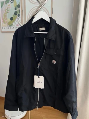 Svart Moncler overshirt med dragkedja - Snygg svart overshirt från Moncler med klassisk krage, bröstficka och logotyp på vänster sida. Jackan har hel dragkedja framtill och är tillverkad i ett lätt, slätt material som passar perfekt för lager-på-lager. Enkel och stilren design. Perfekt för våren/sommaren. Passar 182-187