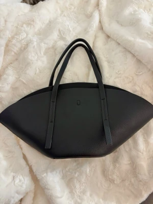 Svart handväska ifrån Gina Tricot  - Snygg svart handväska med unik, bågformad design och långa dubbla axelband. Väskan är tillverkad i ett slätt material som liknar läder och har minimalistiska detaljer. Perfekt för dig som gillar stilrena accessoarer med modern touch. Populär från Gina Tricot 