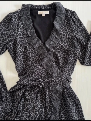 Leopardmönstrad omlottklänning Michael Kors - Snygg leopardmönstrad omlottklänning från Michael Kors i svart och grått. Klänningen har volangkant längs ringningen, knytband i midjan och långa ärmar. Perfekt för dig som vill sticka ut med ett djurmönster och gillar trendiga detaljer.