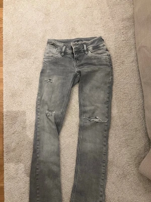 Grå slitna jeans från LTB - Säljer ett par grå jeans från LTB med slitningar på låren och knäna. Jeansen har fem fickor, dragkedjedetalj på myntfickan och raka ben. Perfekta för en avslappnad och trendig look.