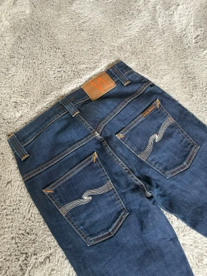 Blå jeans från Nudie Jeans - Säljer ett par klassiska blå jeans från Nudie Jeans med raka ben och snygga kontrastsömmar. Bakfickorna har Nudies ikoniska vågmönster i vitt och läderpatch i midjan. Perfekta för dig som gillar stilrena och tidlösa jeans i mjukt denimtyg.
