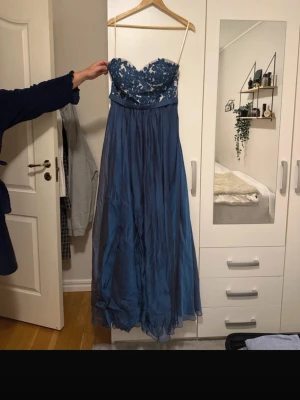 Blå långklänning med spetsdetaljer - Elegant blå långklänning med axelbandslös design och vacker spets upptill. Kjolen är i ett tunt, svepande material som ger en lyxig känsla. Perfekt för bal eller festliga tillfällen. Spetsen upptill har blommönster och klänningen har en markerad midja.