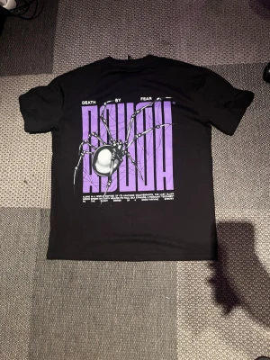 Svart t-shirt med lila tryck och spindel - Cool svart t-shirt med stort lila tryck och spindelgrafik på ryggen samt mindre tryck fram. Texten 'DEATH BY FEAR' och 'ARACHNIA' ger en edgy vibe. Perfekt för dig som gillar streetwear och unika prints.