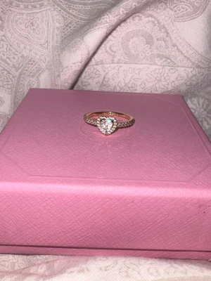 Guldfärgad ring med hjärtformad sten - Snygg guldfärgad Pandora ring med en stor hjärtformad klar sten i mitten och små gnistrande stenar längs bandet. Pris kan diskuteras.