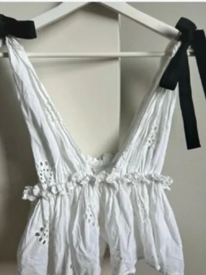 Vit broderad topp med svarta rosetter - Söt vit topp med broderade detaljer och volanger. Axelbanden knyts med svarta rosetter vilket ger en kontrast till den vita bomullen. Djup urringning och luftig passform, perfekt för sommaren.