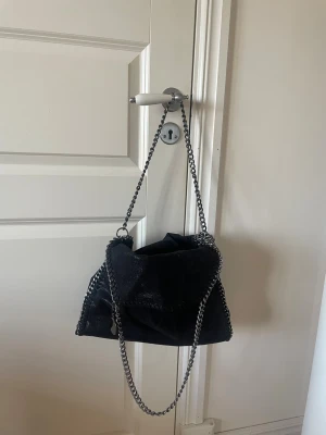 Svart handväska med kedjedetaljer - Cool svart handväska med silvriga kedjor som axelrem och runt kanterna. Väskan har en mjuk, lite slouchy form och är tillverkad i ett material som ser ut som mocka eller sammet. Perfekt för dig som gillar en edgy och trendig look.