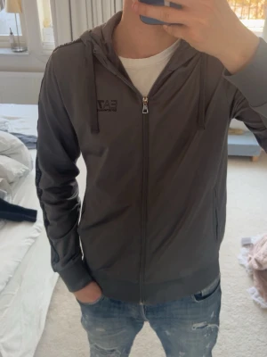 Grå EA7 Emporio Armani hoodie - Snygg grå hoodie från EA7 Emporio Armani med dragkedja framtill och huva med dragsko. Svarta detaljer längs ärmarna med Emporio Armani-logga. Tillverkad i ett mjukt och bekvämt material, perfekt för en avslappnad stil.