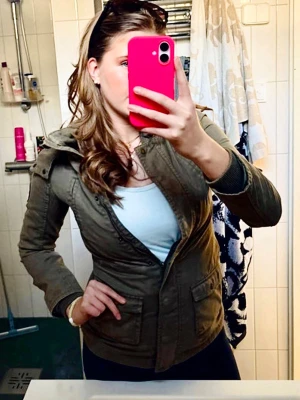 Olivgrön vindjacka från BEL - Snygg olivgrön vindjacka från BEL med huva och dragkedja. Jackan har två stora fickor framtill med knappar, ribbade muddar vid ärmsluten och ett mönstrat innerfoder. Märkespatch på ärmen ger extra stilpoäng. Perfekt för blåsiga dagar.