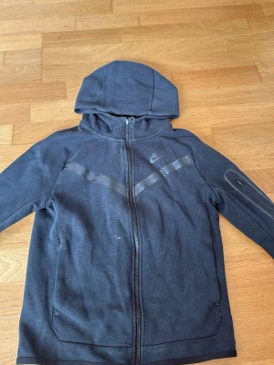 Svart hoodie från Nike med dragkedja - Snygg svart hoodie från Nike med hel dragkedja framtill och huva. Hoodien har diskreta reflexdetaljer över bröstet och en ficka med dragkedja på ena ärmen. Tillverkad i mjukt bomullsmaterial, perfekt för en sportig och avslappnad stil.