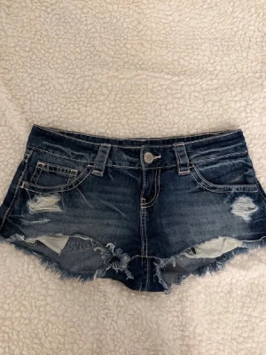 Blå slitna jeansshorts med fransar - Säljer ett par blå jeansshorts med slitna detaljer och fransig kant. Shortsen har fem fickor, låg midja och coola sömmar på bakfickorna. Perfekta för sommaren och ger en avslappnad vibe.