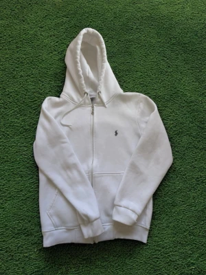 Vit zip hoodie från polo Ralph Lauren - Snygg vit hoodie med dragkedja från polo Ralph Lauren. Klassisk design med huva och dragsko, samt fickor framtill. Liten broderad logga på bröstet. Perfekt för en clean och stilren look. Tillverkad i mjukt bomullsmaterial. Storlek xl men passar till medium också