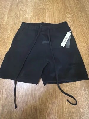 Svarta shorts från Essentials - Säljer ett par svarta shorts från Essentials med långa dragsnören i midjan och diskret logga framtill. Shortsen har en avslappnad passform och är tillverkade i mjukt sweatshirtmaterial. Perfekta för en chill och trendig look.