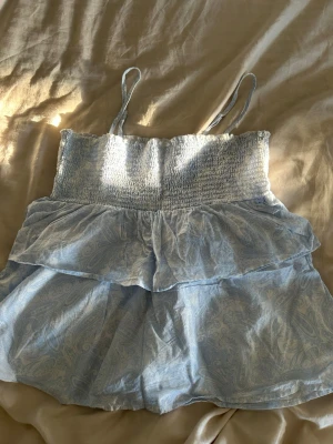 Ljusblå topp med volanger Gina Tricot - Supersöt ljusblå topp från Gina Tricot med tunna axelband och smock upptill. Toppen har dubbla volanger och ett diskret paisleymönster i tyget. Perfekt för sommardagar och enkel att matcha med jeans eller kjol. Använt en gång och är köpt på young avdelning 