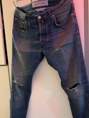 Blå slitna jeans från Jacob Cohën - Säljer ett par blå jeans från Jacob Cohën med snygga slitningar och hål på benen. Jeansen har klassisk femficksmodell, knappgylf och är tillverkade i mjukt denimtyg. Perfekta för en avslappnad och trendig look. Har en defekt där en av fickorna är spruckna och därför lägger jag det på ett lägre pris