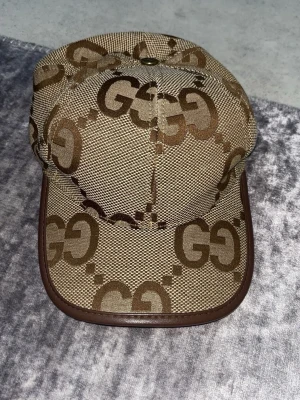 Beige GG-mönstrad keps från Gucci - Säljer en beige keps från Gucci med det klassiska GG-mönstret i brunt över hela kepsen. Kepsen har böjd skärm med brun kant och är tillverkad i ett slitstarkt tygmaterial. Perfekt för dig som vill ha en stilren och lyxig accessoar.