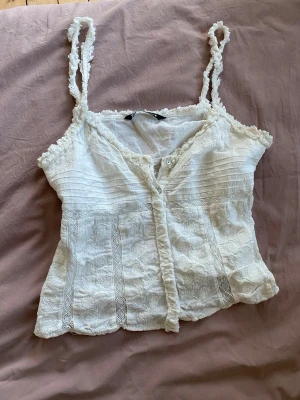 Vit spetsig linnetopp från Zara - Supersöt vit linnetopp från Zara med spetsdetaljer, knappar framtill och smala axelband. Toppen har romantiska volanger och broderade partier som ger en bohemisk känsla. Perfekt att styla med jeans eller kjol för en trendig look.