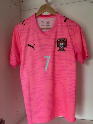 Rosa Portugal fotbollströja Puma - Säljer en rosa Portugal fotbollströja från Puma med nummer 7 och namnet Ronaldo på ryggen. Tröjan har korta ärmar, rund hals och Portugal-emblemet på bröstet. Perfekt för dig som älskar fotboll och vill sticka ut på planen.