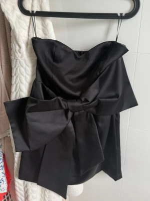 Svart axelbandslös klänning med rosett - Supersnygg svart axelbandslös klänning med stor rosett framtill. Klänningen är kort och har en elegant, glansig finish som ger en lyxig känsla. Perfekt för dig som vill sticka ut med en unik detalj. Materialet känns som satin.