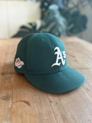 Grön Oakland Athletics keps - Mörkgrön keps från New Era med Oakland Athletics 'A's' broderat i vitt framtill och en patch på sidan. Klassisk basebollmodell med böjd skärm och broderade ventilationshål. Perfekt för dig som gillar sportig streetstyle. Storlek 57,7 cm
