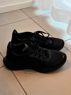 Svarta Adidas sneakers - Snygga helsvarta sneakers från Adidas helt oanvända, med klassisk låg profil och diskret logga på hälen. Skorna har ovandel i mesh och syntet, samt matchande svarta snören och sula. Perfekta för dig som gillar stilrena och sportiga skor. Storlek 39/5 