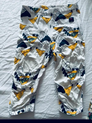 Färgglada leggings från Polarn O. Pyret - Säljer ett par vita leggings från Polarn O. Pyret med coolt tryck av flygande ögon och gula trumpeter. Mjukt och stretchigt material som passar perfekt för lek och bus. Elastisk midja för skön passform. Storlek 98, för barn 2-3 år.