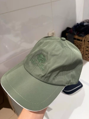 Olivgrön keps från Loro Piana - Snygg olivgrön keps från Loro Piana med broderad logga och emblem framtill. Klassisk böjd skärm och justerbar passform bak. Tillverkad i ett slätt, tåligt material som ger en clean och stilren look.