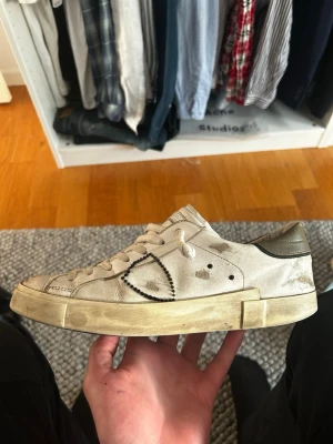 Philippe Model vita sneakers - Säljer ett par vita Philippe Model sneakers med beige och olivgröna detaljer. Skorna har en låg silhuett, klassisk snörning och är tillverkade i läder med distinkta sömmar och logotyp på sidan. Sulan är beige och ger en vintagekänsla. Perfekta för en avslappnad streetwear-look.