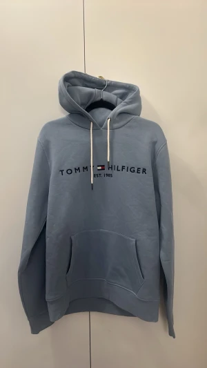 Ljusblå hoodie från Tommy Hilfiger - Ljusblå hoodie från Tommy Hilfiger med broderad logga och text 'EST. 1985' på bröstet. Klassisk känguruficka framtill, vita dragsnören och mjukt material. Perfekt för en avslappnad och stilren look. Får kläder av bonus pappa för att sälja eftersom han inte använder de.