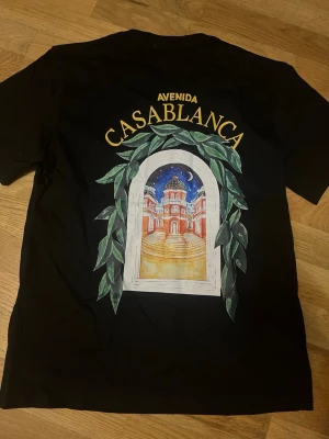 Svart t-shirt från Casablanca - Svart t-shirt från Casablanca med färgglatt tryck på ryggen som föreställer en byggnad och texten 'Avenida Casablanca'. Klassisk passform och rund hals. Perfekt för dig som gillar unika prints och streetwear vibes.