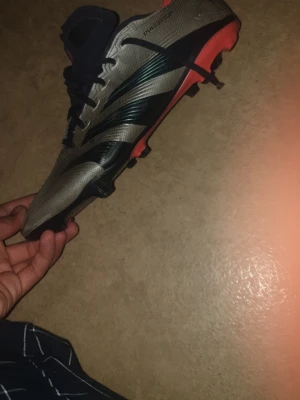 Adidas Predator fotbollsskor grå/svart - Säljer ett par Adidas Predator fotbollsskor i grått med svarta och mörkblå detaljer samt orange sula. Skorna har snörning och är designade för bra grepp på planen. Snygg och modern look med klassiska Predator-linjer.