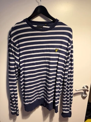 Randig långärmad tröja Lyle & Scott - Hej säljer en Snygg långärmad tröja från Lyle & Scott med marinblå och vita horisontella ränder. Tröjan har rund halsringning, ribbade muddar och den klassiska gula loggan på bröstet. Perfekt för en avslappnad och stilren look.