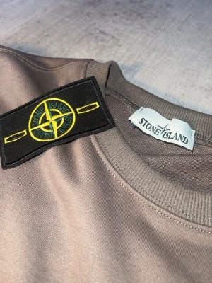 Beige sweatshirt från Stone Island - Snygg beige sweatshirt från Stone Island med klassisk rund hals och ikonisk svart patch med gult och grönt kompassmärke på bröstet. Tröjan har ribbade muddar vid ärmslut och nederkant. Perfekt för en stilren och avslappnad look.
